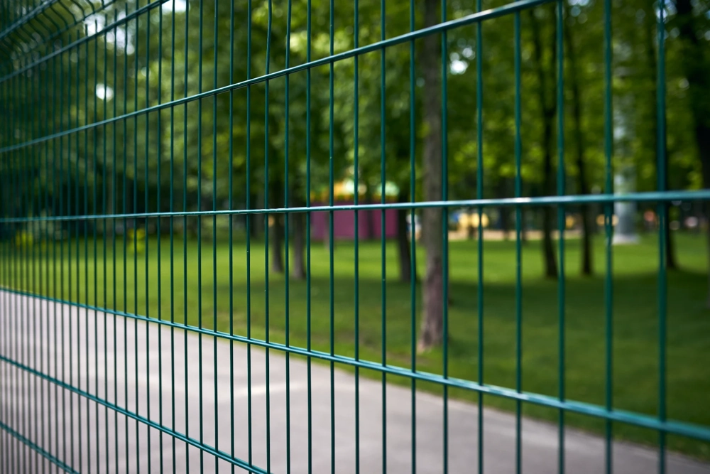 Ogrodzenia panelowe 2d: zielone metalowe przęsło ogrodzenia z płaskich prętów przy chodniku, widoczne słupki